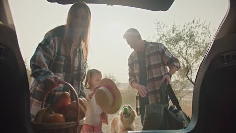 a middle-aged man with gray hair in a plaid shirt and his wife, a brunette girl in a green plaid shirt, together with their daughter, are packing things that are necessary for a picnic while driving back home after a walk in the summer