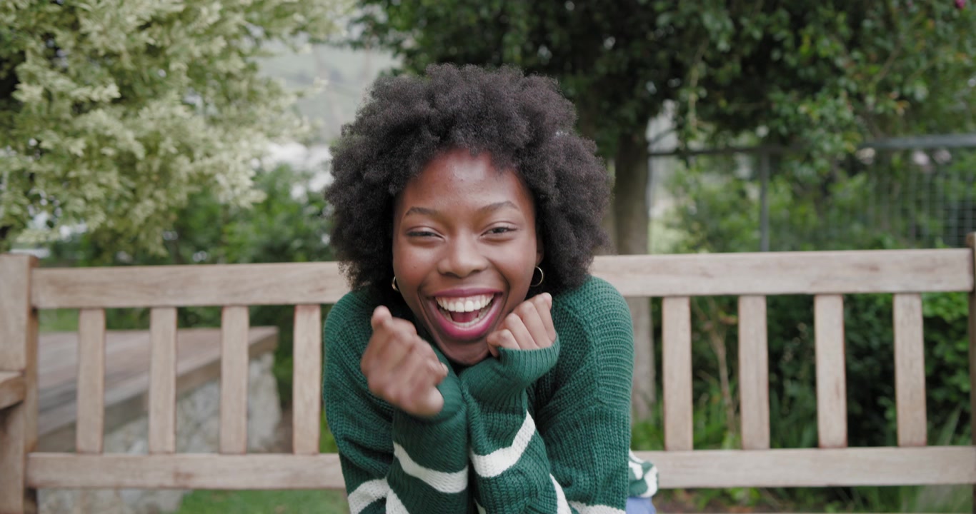 Femme afro-américaine d'une vingtaine d'années assise sur un banc en bois dans un parc, observant et riant.