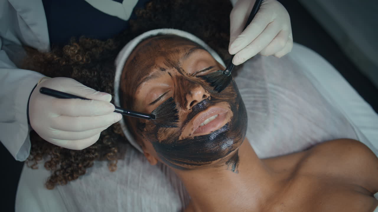 Primo piano di estetista che applica maschera con pennello per peeling in salone. Bella modella vista dall'alto