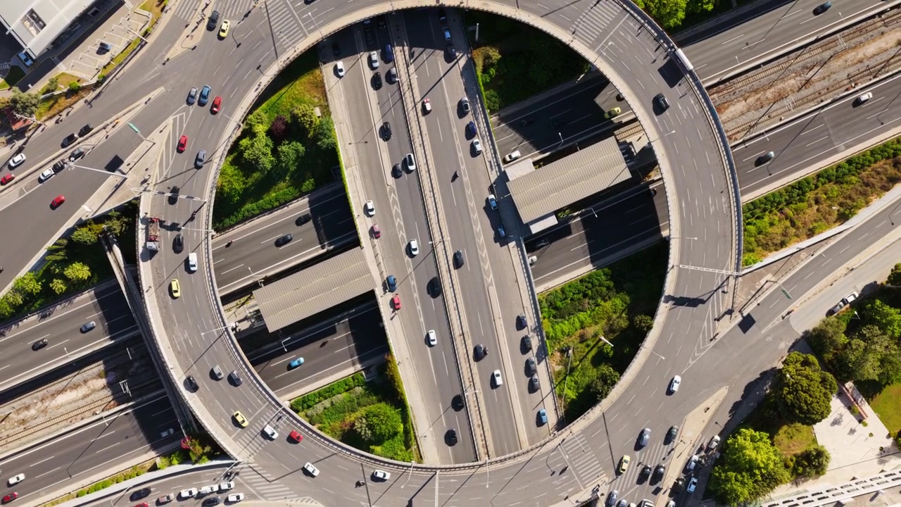 Rising aerial top-down view of a complex highway interchange with a large roundabout, showcasing cars and vehicles moving through the intricate road network and infrastructure on a sunny day