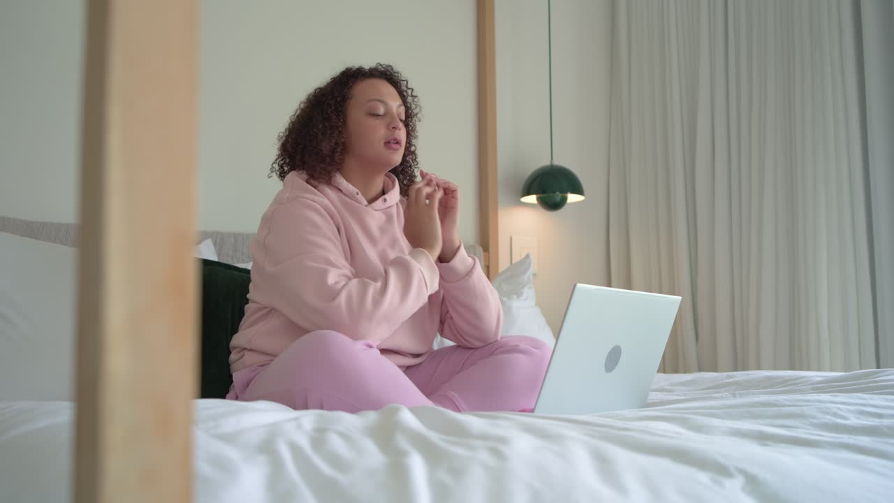 Donna afroamericana adulta con felpa rosa che digita sul letto, si ferma, riflette e riprende a lavorare sul laptop