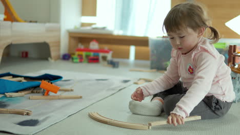 cute toddler girl building wooden railway track connecting pieces sitting on a floor in playroom at home