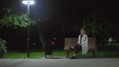 lady seated alone on wooden bench under glowing streetlamp at night, holding phone in hand while gazing into distant space with tree leaves swaying in wind and red light glowing in background