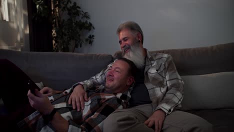 A-happy-brunette-man-in-a-checkered-shirt-lies-on-his-elderly-boyfriend-with-gray-hair-and-a-lush-and-white-beard-in-a-checkered-shirt-and-they-watch-TV-while-sitting-on-a-gray-sofa-in-a-modern-apartment