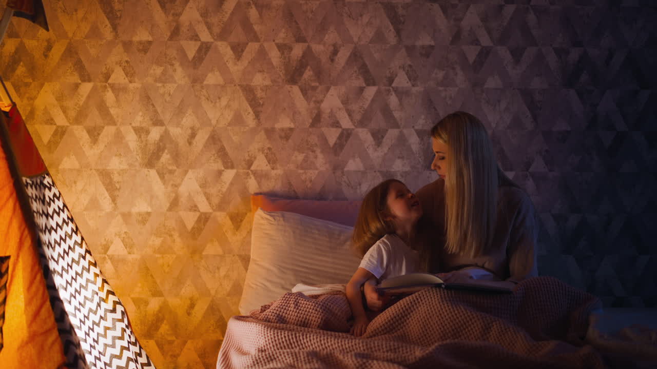 Little girl and mum with book on bed at illuminated wigwam