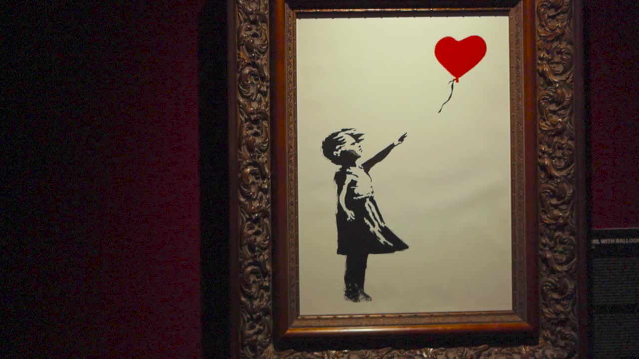 Famous girl with balloon painting done by Banksy at his exhibition in Tallinn Estonia. Shows a little girl letting go of a red heart shaped balloon. Painting is in a golden frame and on a red wall.