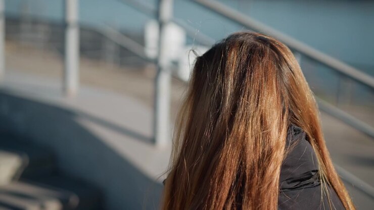 Donna con lunghi capelli castani in piedi all'aperto vicino a uno specchio d'acqua, di spalle alla fotocamera. La scena è illuminata dolcemente dalla luce naturale del giorno, creando caldi riflessi nei suoi capelli e proiettando ombre delicate.