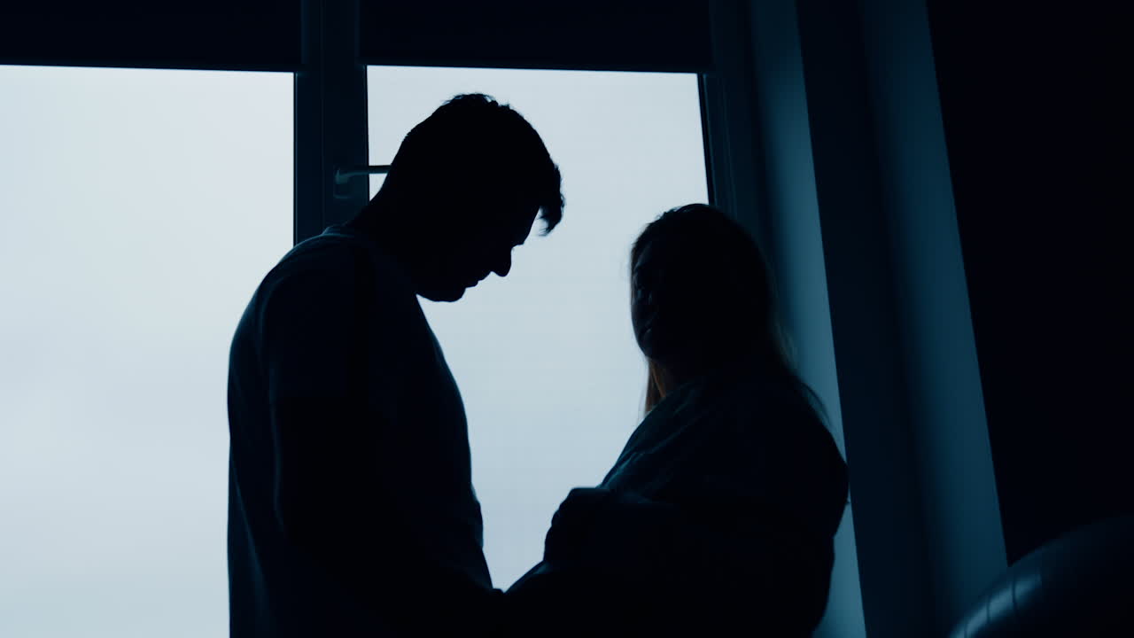 Silhouettes of expecting couple standing at the window. Man strokes big wife's belly and then embraces her. Low angle view.