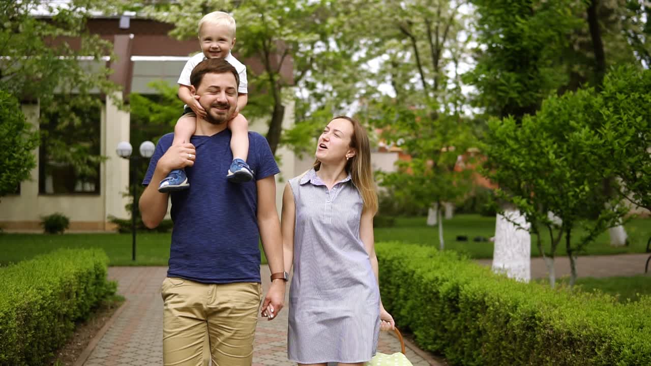 Happy family walking on the street path together on a sunny day. Baby boy is riding on the father's shoulders. Caucasian family. Front view