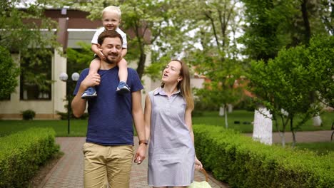 Happy-family-walking-on-the-street-path-together-on-a-sunny-day.-Baby-boy-is-riding-on-the-father's-shoulders.-Caucasian-family.-Front-view