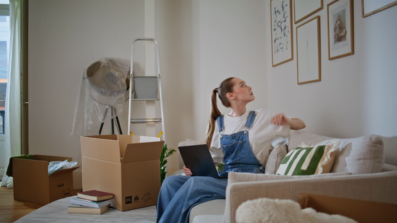 Woman choosing decor online sitting couch with laptop looking on wall new home