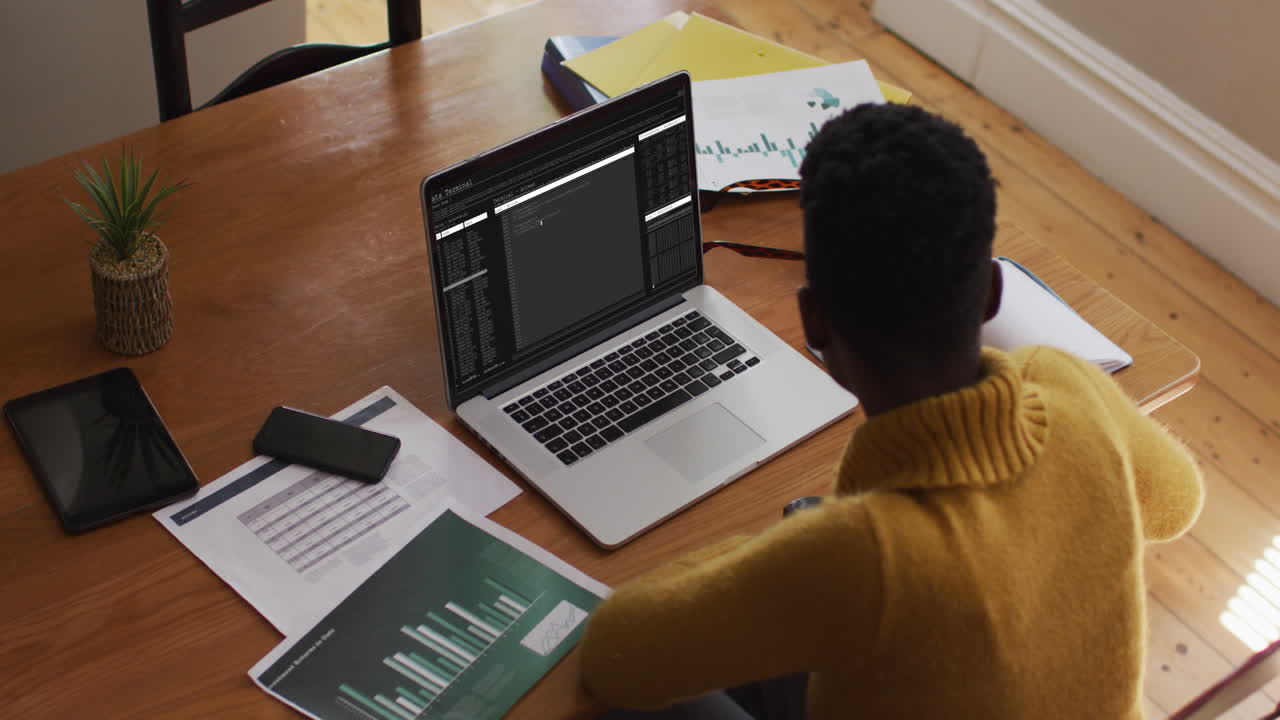 African american man sitting at desk watching coding data processing on laptop screen