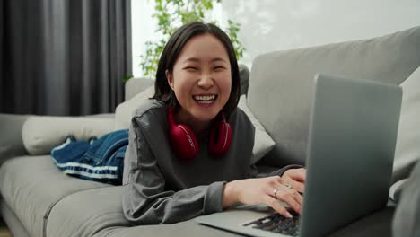 Porträt-Einer-Asiatischen-Frau,-Die-Auf-Dem-Sofa-Liegt,-Auf-Dem-Laptop-Tippt,-In-Die-Kamera-Schaut-Und-Lächelt