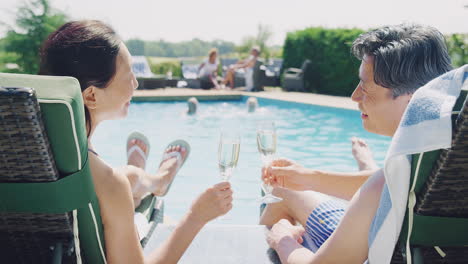 mature couple on loungers relaxing by swimming pool on summer vacation drinking champagne