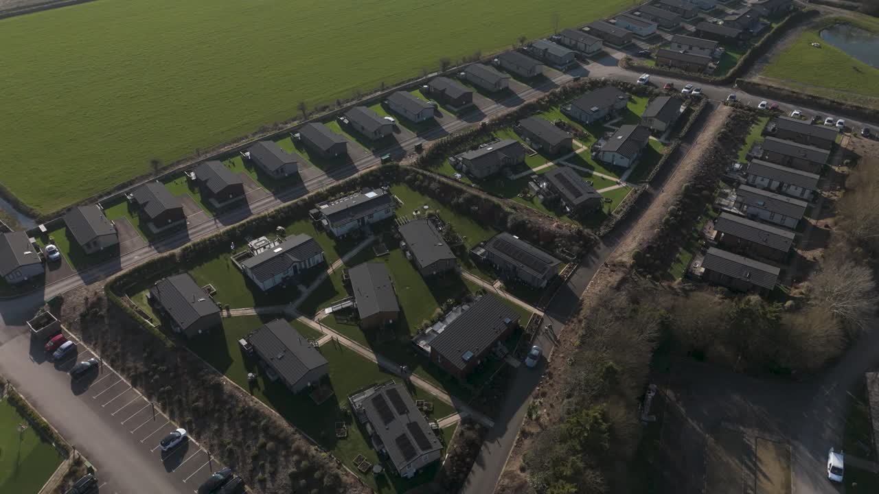Golden-hour aerial of holiday lodge park with curved rows of cabins, warm light and long shadows over neat grass depicting tranquil seasonal getaway