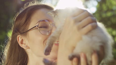 close-up view of a caucasian woman in glasses holding small cat and kissing it in the park on a summer day