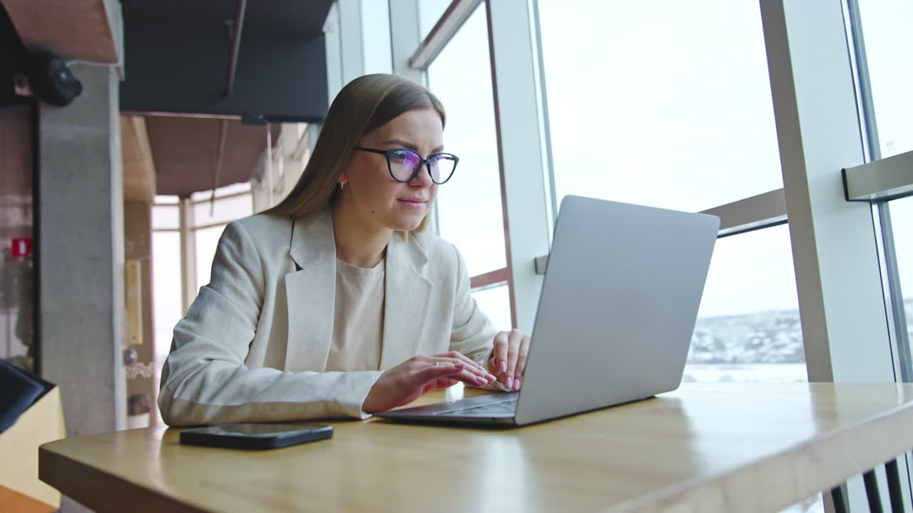 Young woman works on her laptop sitting at the desk. Lady in glasses looking intently at the screen. Workplace at the panoramic window.