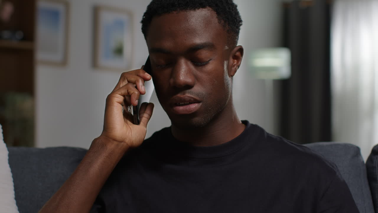Ulykkelig og deprimert ung mann sitter i sofaen hjemme og snakker med en hjelpelinje for støtte på mobiltelefon