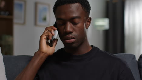 unhappy and depressed young man sitting on sofa at home talking to helpline for support on mobile phone