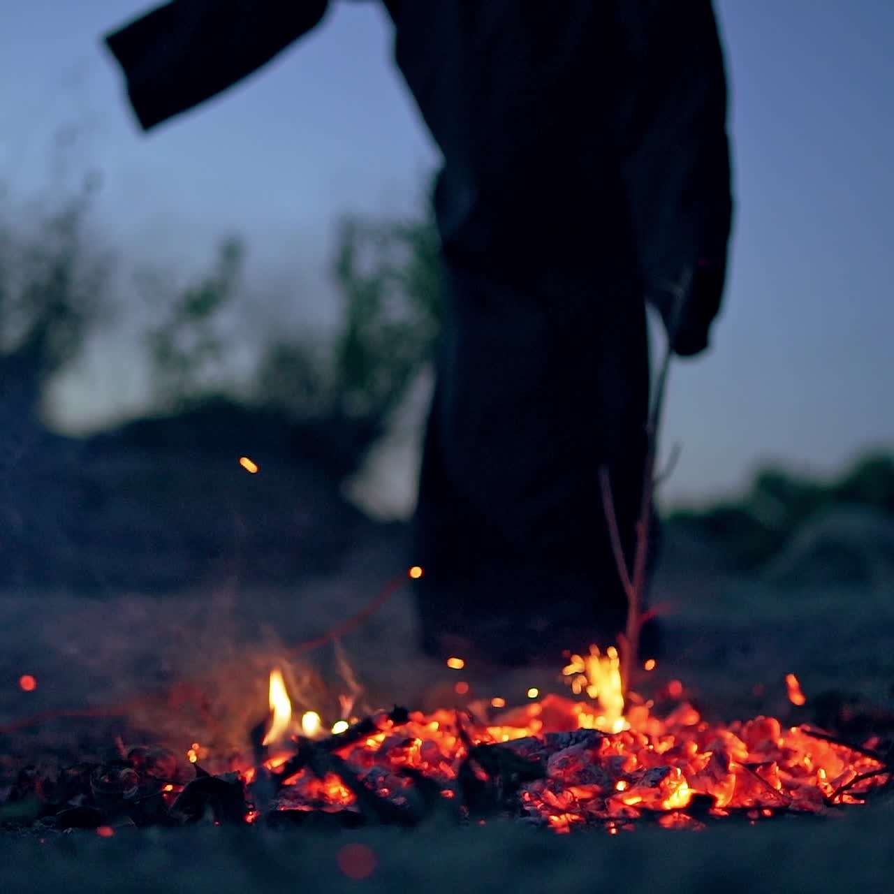 Unrecognizable figure in black is putting out bonfire. Witch with stick splits embers of burnt-out fire outdoors.