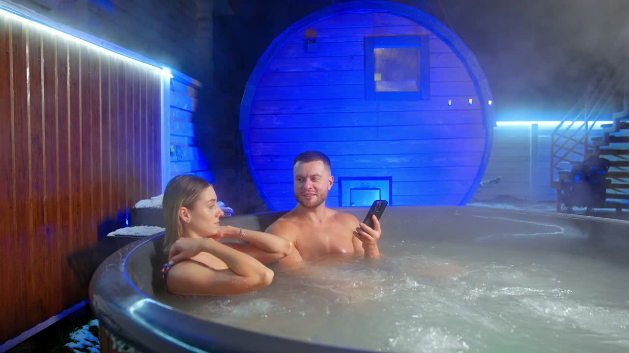 Caucasian family having relaxation in a hot tub in winter. Man is holding a smartphone in his hand.
