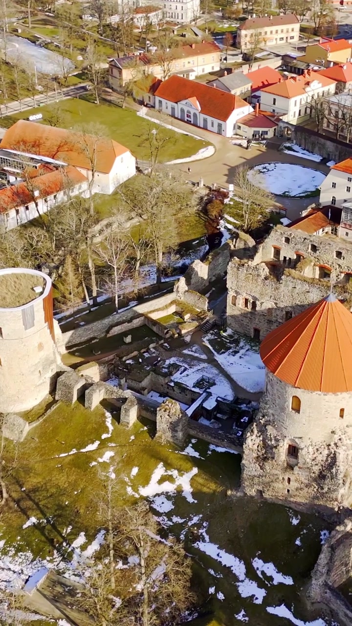 Vertical aerial of a historic fortress complex with round towers, ruined walls, and inner grounds, showing heritage architecture, seasonal textures, and urban surroundings.