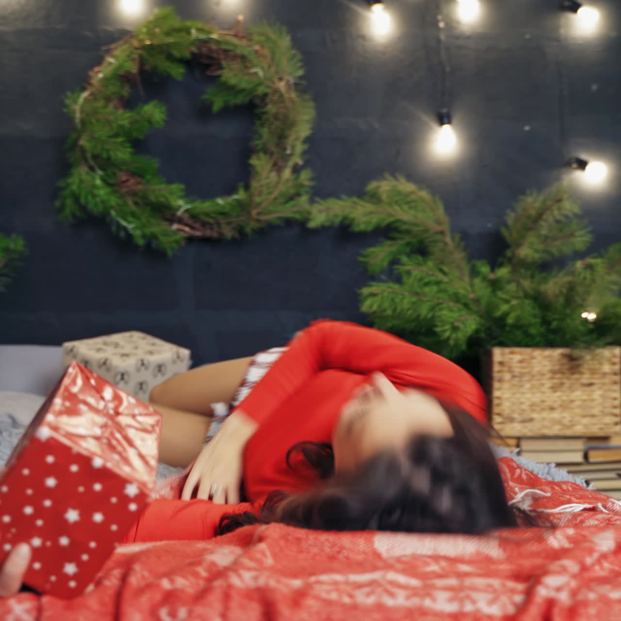 Woman in warm stylish clothes poses on bed in decorated room. Beauty in red sweater happy to receive gifts. Fest spirit