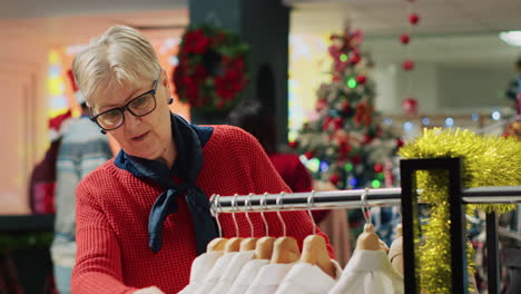 elderly woman browsing through clothes in xmas adorn clothing store during winter holiday season. senior client in shopping spree session at christmas decorated fashion boutique in mall