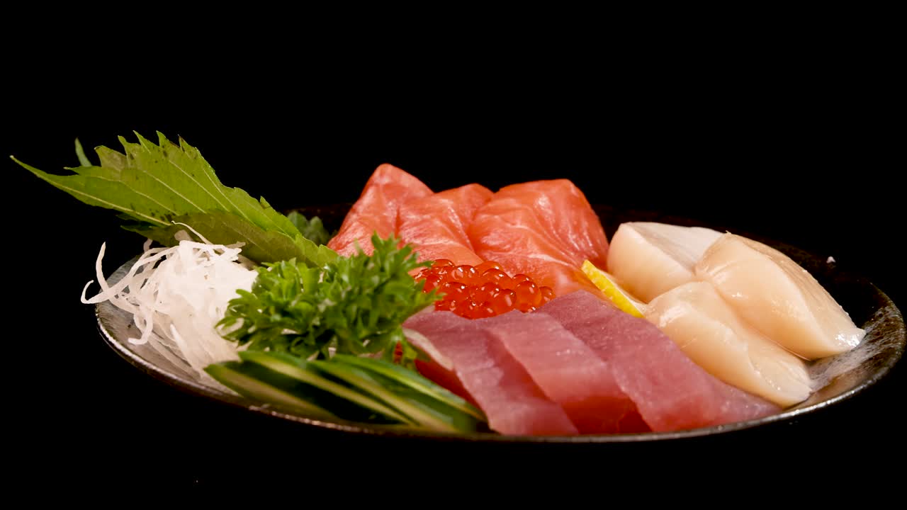 A plate of fresh sashimi, including salmon, tuna, scallop, and salmon roe, rotates smoothly under studio lighting against a black background