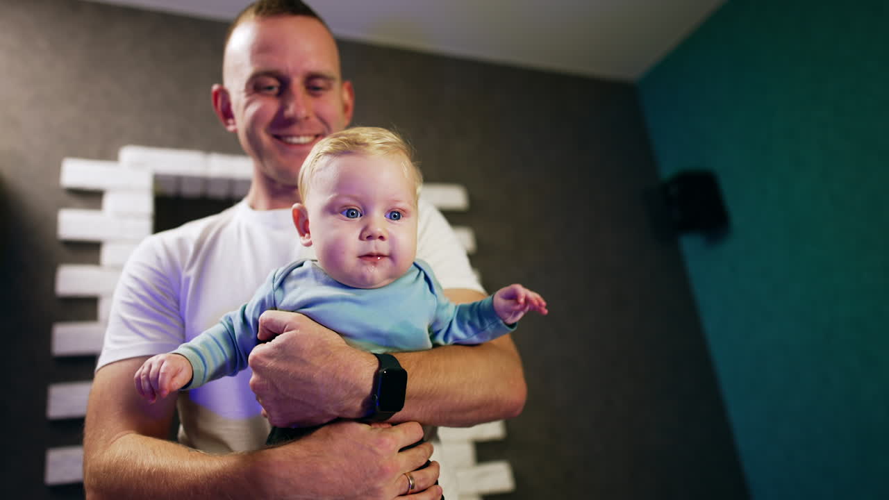 Happy smiling Caucasian man holding a baby in front of him. Cute plump kid looks at something smiling adorably.