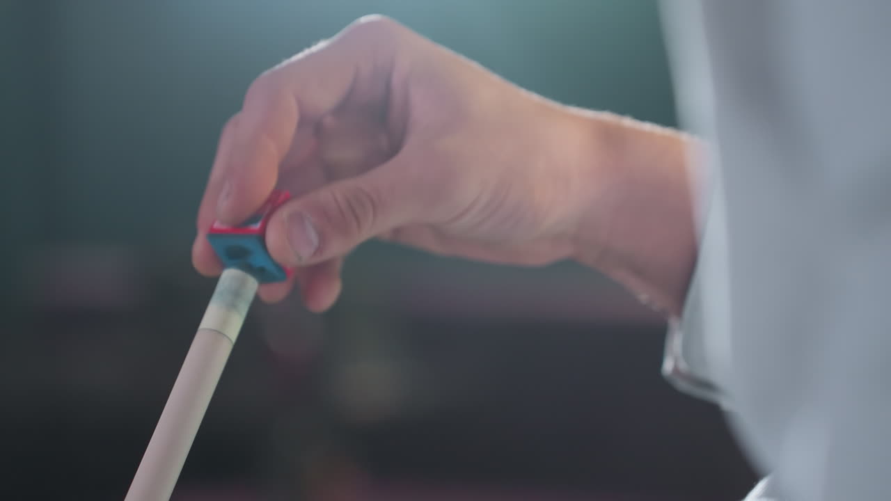 toma de primer plano de la mano con camisa blanca aplicando tiza roja y azul al palo de señal antes del juego de billar. el agarre enfocado y el fondo borroso enfatizan la preparación para el siguiente movimiento