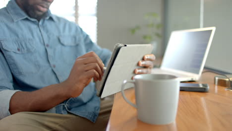 using stylus on tablet, african american man working from home with laptop and coffee