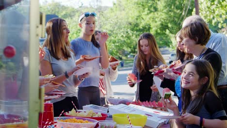 neighbours stand eating around a table at a block party