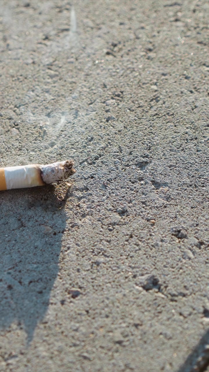 Primer plano de un cigarrillo quemado reposando sobre suelo de hormigón rugoso con iluminación suave. La ceniza y la marca de quemadura son claramente visibles, enfatizando los detalles de textura tanto en el material de fumar como en el suelo.