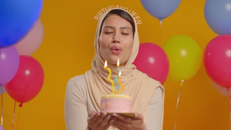 studio portrait of woman wearing hijab and birthday queen headband celebrating birthday blowing out candles on cake