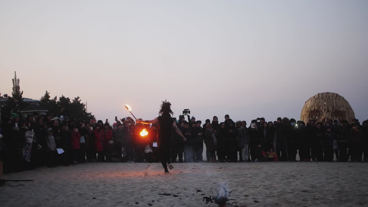 Slow motion clip of a male stunt performer performing fire dance using torches on a spinning rod during fire festival on a beach in South Korea at sunset.