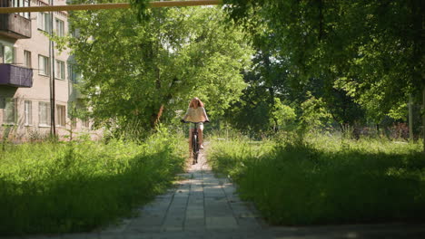 young woman confidently rides bicycle along a sunlit, lush green park path surrounded by tall grass and leafy trees, with her long hair flowing freely, with a building in the background