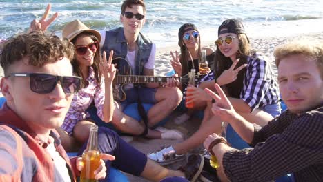 Young-man-recording-a-video-or-taking-selfie-of-group-of-friends-sitting-on-easychairs-on-the-beach,-playing-guitar-and-singing-on-a-summer-evening.-Slowmotion-shot