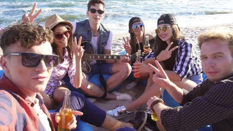 young man recording a video or taking selfie of group of friends sitting on easychairs on the beach, playing guitar and singing on a summer evening. slowmotion shot