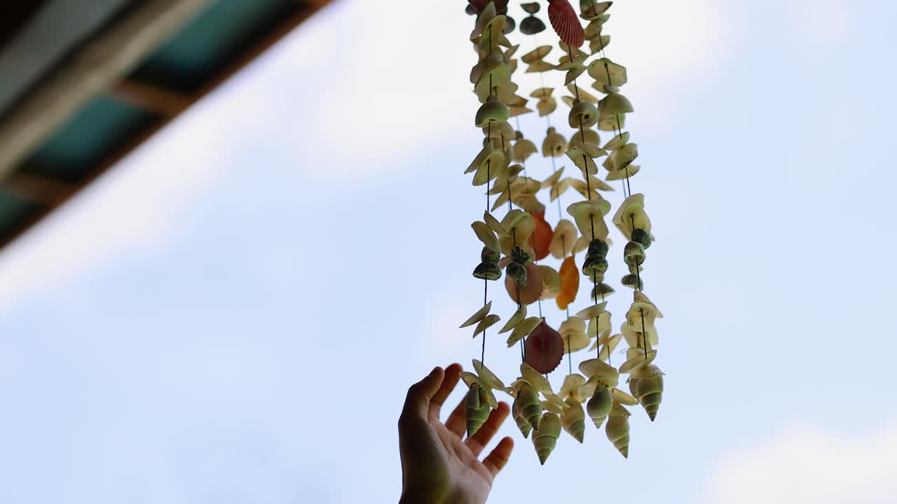 A hand gently interacts with a hanging shell garland against a bright sky in Phuket, Thailand. Natural lighting enhances the serene atmosphere