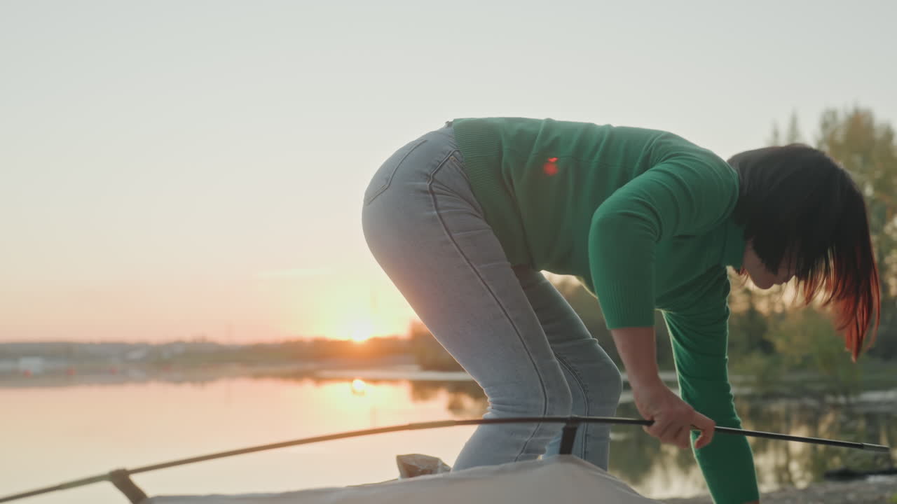 Woman setting up lakeside tent at sunset, green sweater and jeans, assembling tent body on pebble shore, calm water reflection, tree line and warm twilight light, quiet solo camping moment