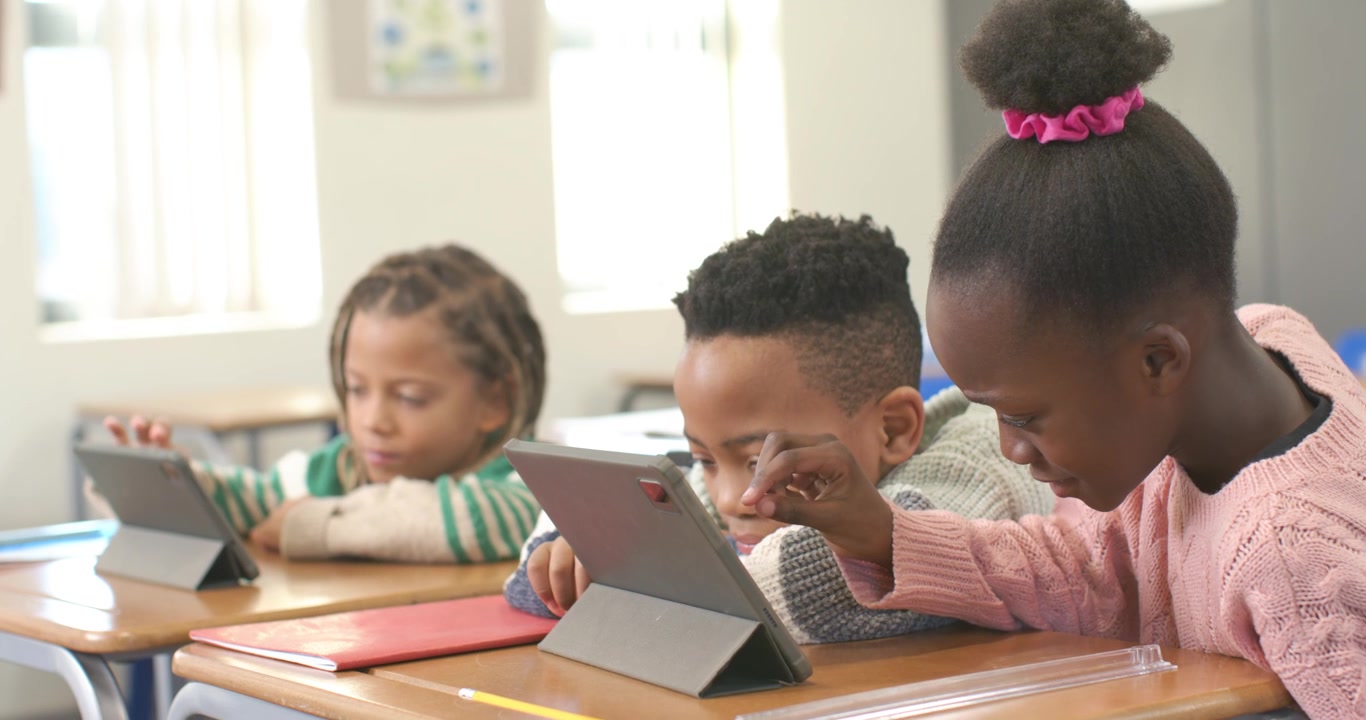 Tre bambini a scuola, una bambina si allunga e tocca un tablet centrale per aiutare un bambino che si china e segue.
