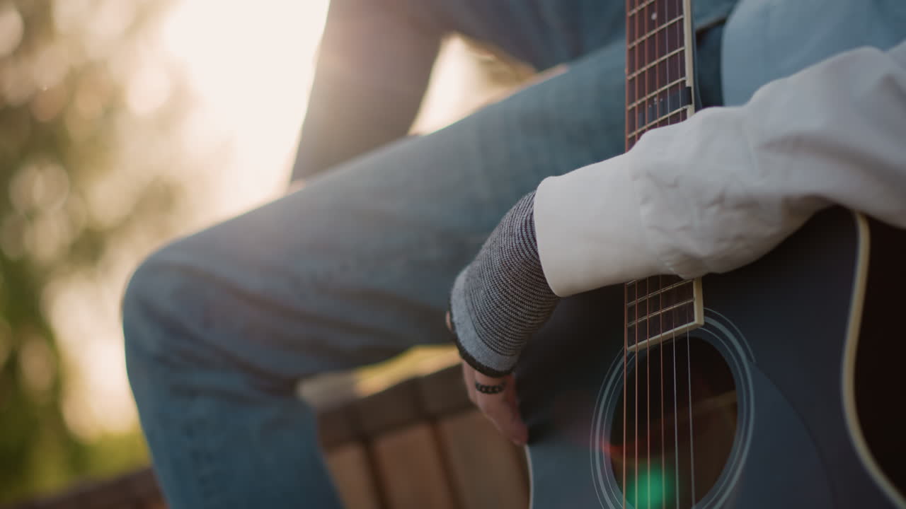 chitarrista bianco che pizzica una chitarra acustica con una mano guantata, primo piano del corpo e del polso, retroilluminazione calda, energia spontanea da artista di strada, ritmo dolce, ambientazione su una panchina del parco, attenzione agli accordi e alla mano
