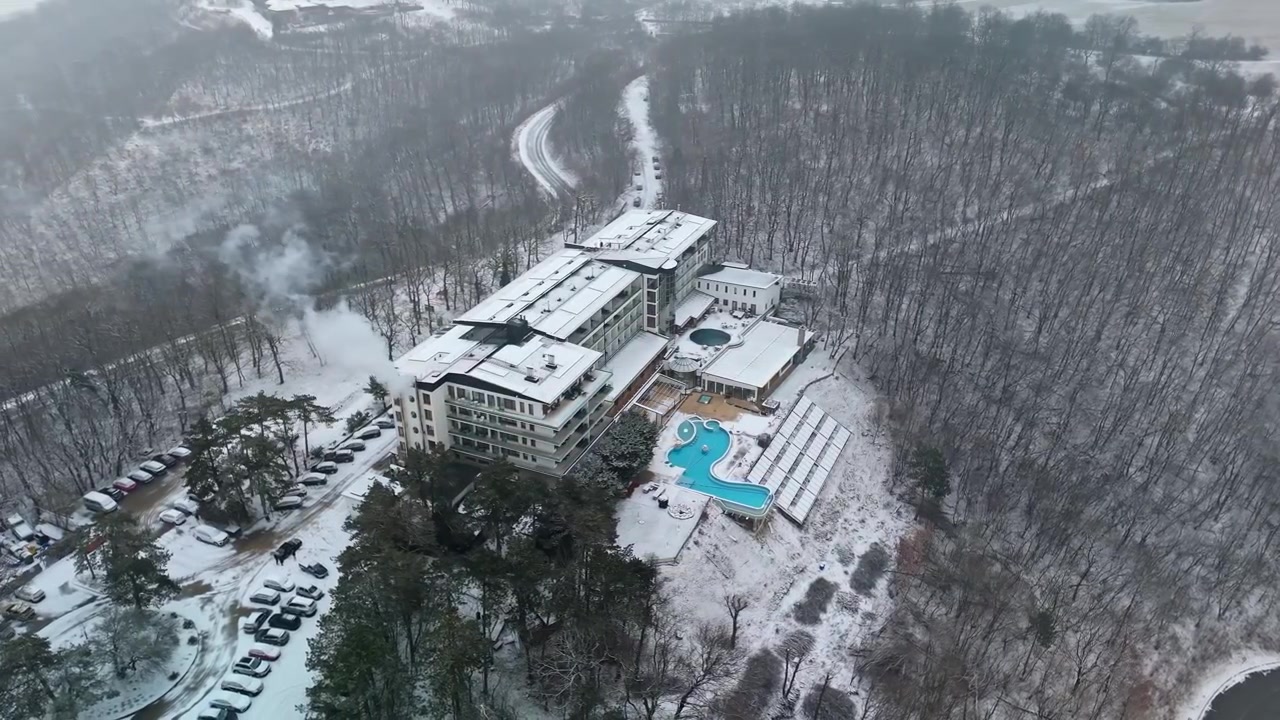 V maďarských Visegrádských horách se uprostřed zasněženého lesa nachází moderní horský hotel s pokoji s balkonem, poblíž termálních lázní a venkovního bazénu, viděno z ptačí perspektivy v zimě.
