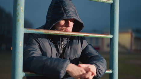 close-up of a person in a hooded jacket leaning on an iron bar with a blur view of a yellow painted building in the background