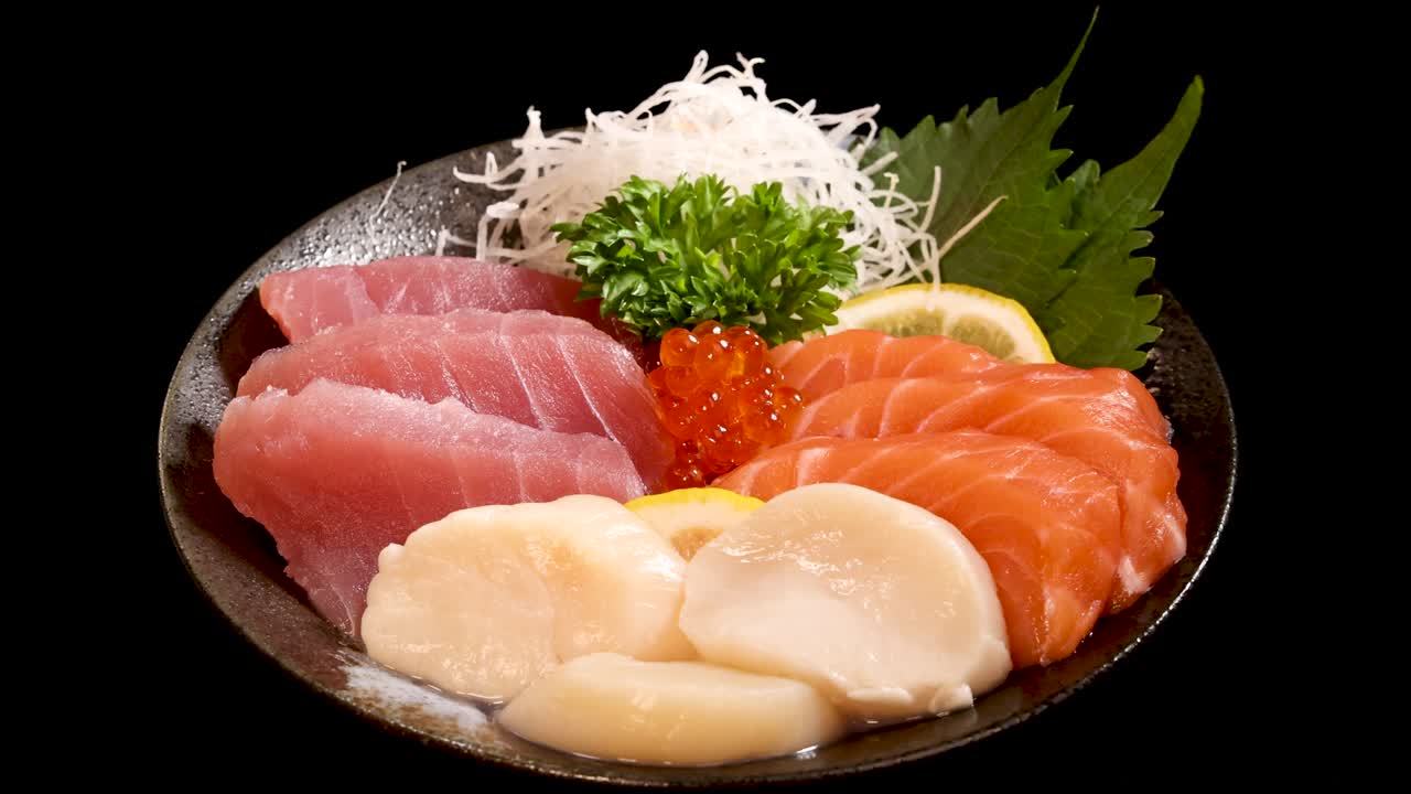A rotating close-up of a Japanese sashimi platter with salmon, tuna, scallop, salmon roe, and garnishes, set against a black background under bright studio lighting