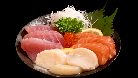 a rotating close-up of a japanese sashimi platter with salmon, tuna, scallop, salmon roe, and garnishes, set against a black background under bright studio lighting