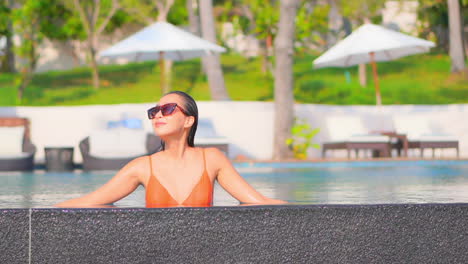 Rich-Sexy-American-Woman-Relaxing-in-Luxury-Hotel-Lounge-Inside-Swimming-Pool-Leaning-on-the-Border,-Line-of-Umbrellas-and-Deckchairs-on-background,-front-view-slow-motion