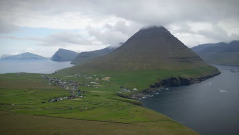 capturing a dramatic aerial view of tviðareiði, faroe islands, revealing a remote village nestled between the ocean and towering, cloud-shrouded mountains