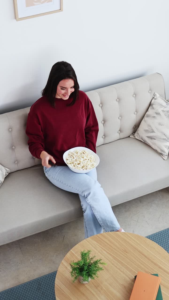 Ung kvinde slapper af på sofaen, spiser popcorn og ser tv